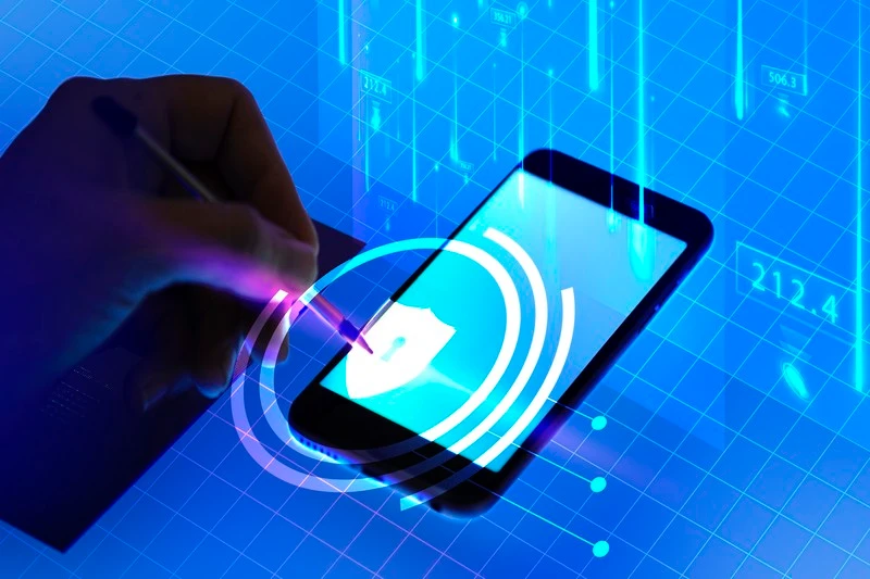 A hand uses a stylus on a glowing smartphone screen with a shield icon, symbolizing cybersecurity. The background shows digital data and grids.