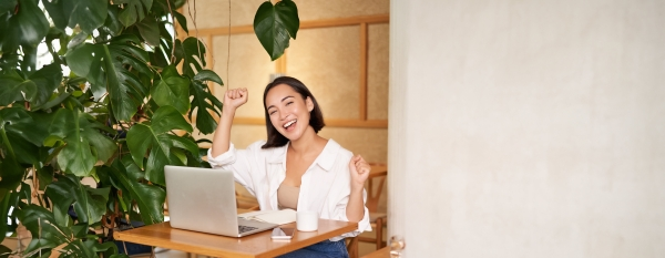 Woman sitting at a desk with a laptop and coffee cup, smiling and raising her arms in excitement, next to a large leafy plant.